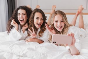 Three european adorable women 20s wearing white bathrobe lying in luxury bedroom at home and taking selfie photo on mobile phone during bridal shower
