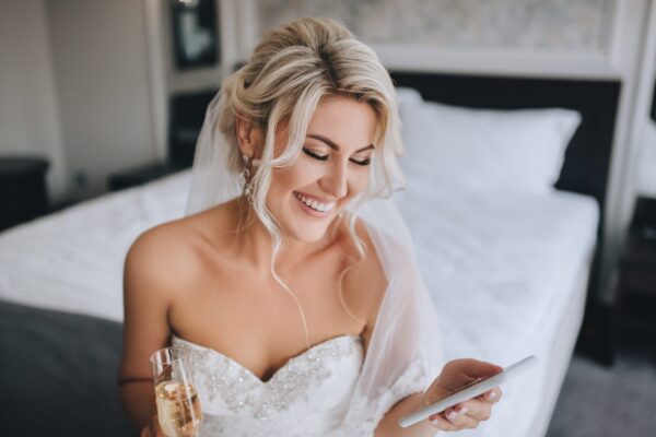 A beautiful, smiling blonde bride in a white dress is sitting on the bed and looking at the phone, reading a message and drinking champagne. Wedding portrait of a cheerful girl.